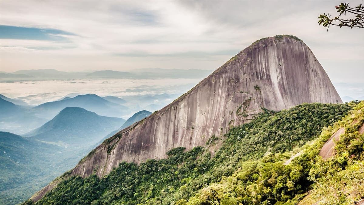 Parede lisa e inclinada do Escalavrado sobre a mata, com mar de nuvens e montanhas da baixada ao fundo, vista do Dedo de Deus